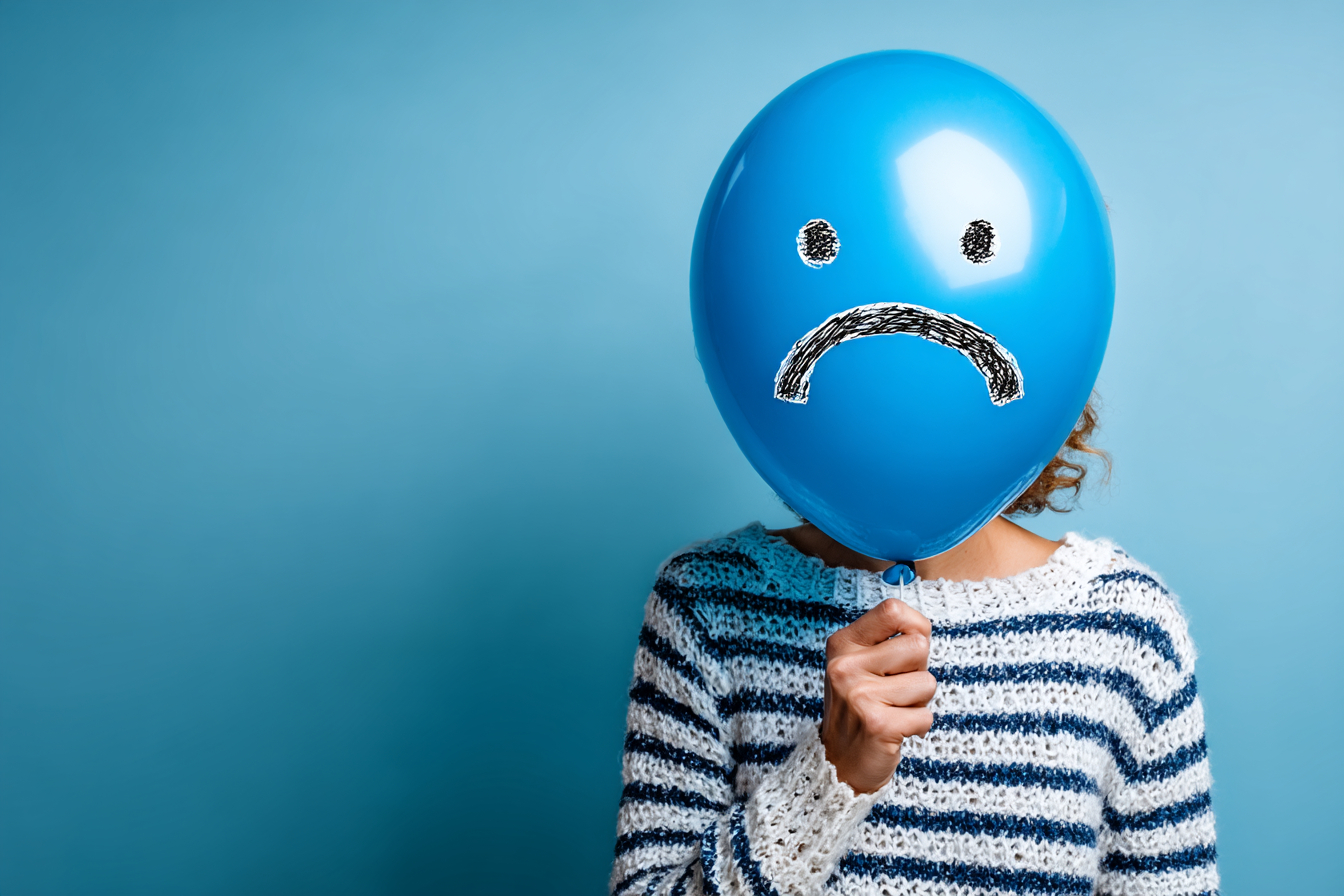A young woman holding a balloon with a sad face against a blue background