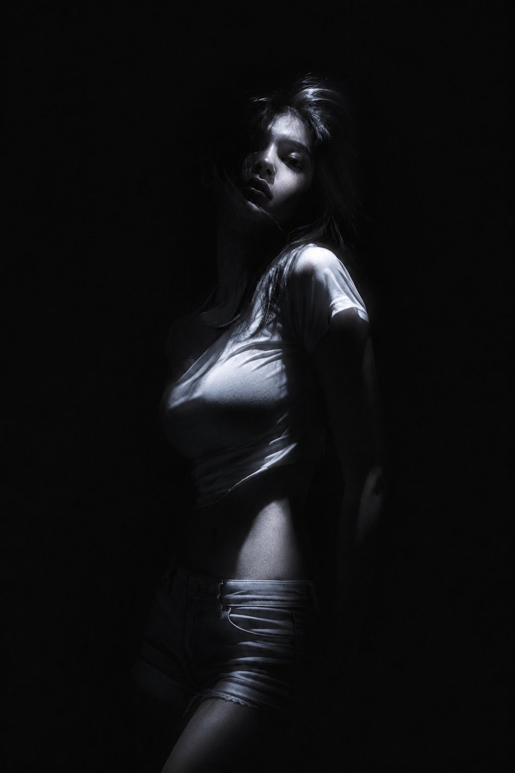 A woman in near-total darkness with a thin light highlighting her features, wearing a loose white tee and low-slung denim shorts, messy hair covering her eyes, looking back over her shoulder with a distant, haunted expression