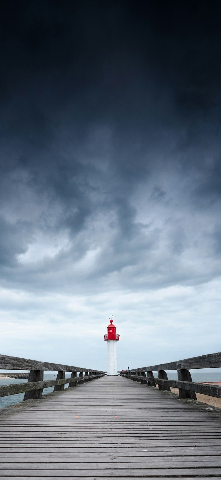 Under a cloudy sky, the scene of the boardwalk and lighthouse is perfect for a phone wallpaper