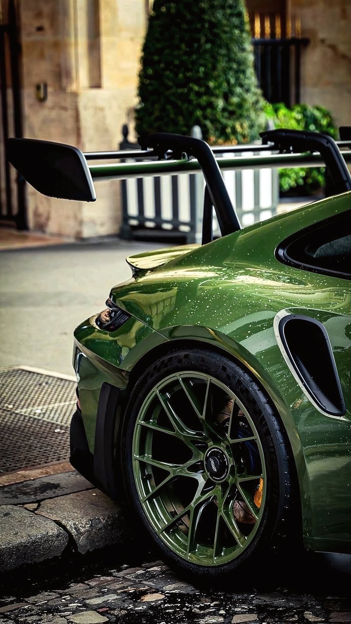 A low-angle shot of an olive green Porsche GT3 RS parked by a cobblestone road with water droplets on its body
