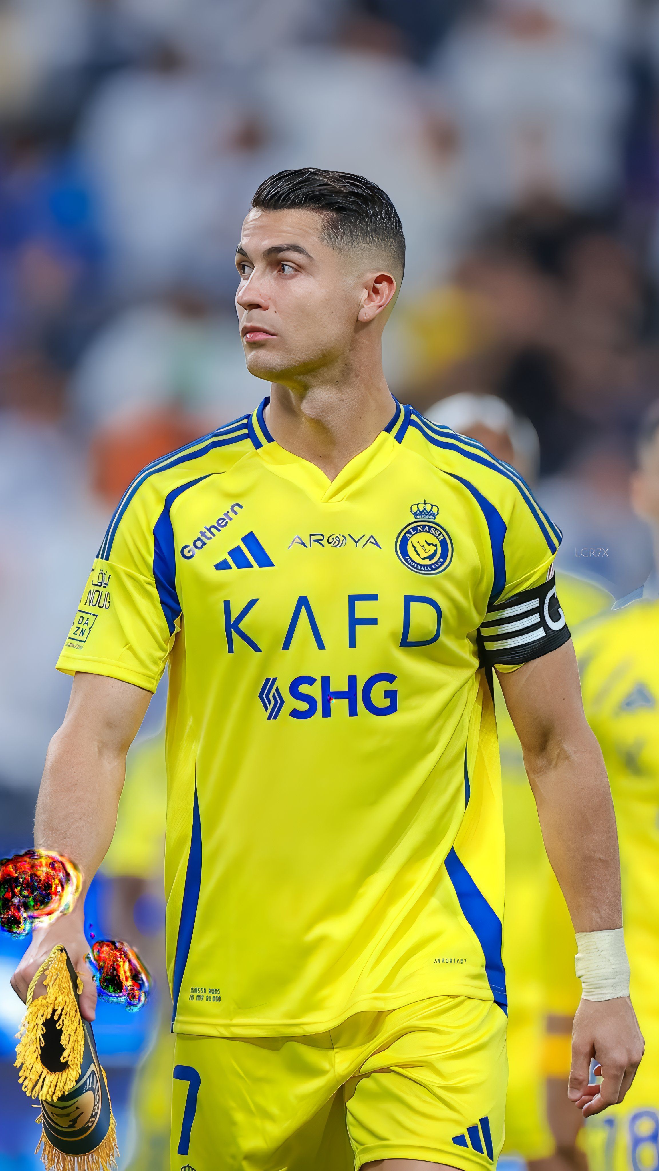 Cristiano Ronaldo vestido con el uniforme amarillo y azul brillante del Al-Nassr sosteniendo una cinta de capitán y un trofeo