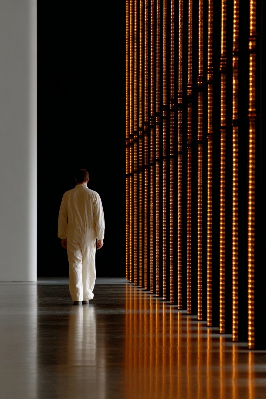 A figure in a white jumpsuit walks towards a towering wall of amber LED strips