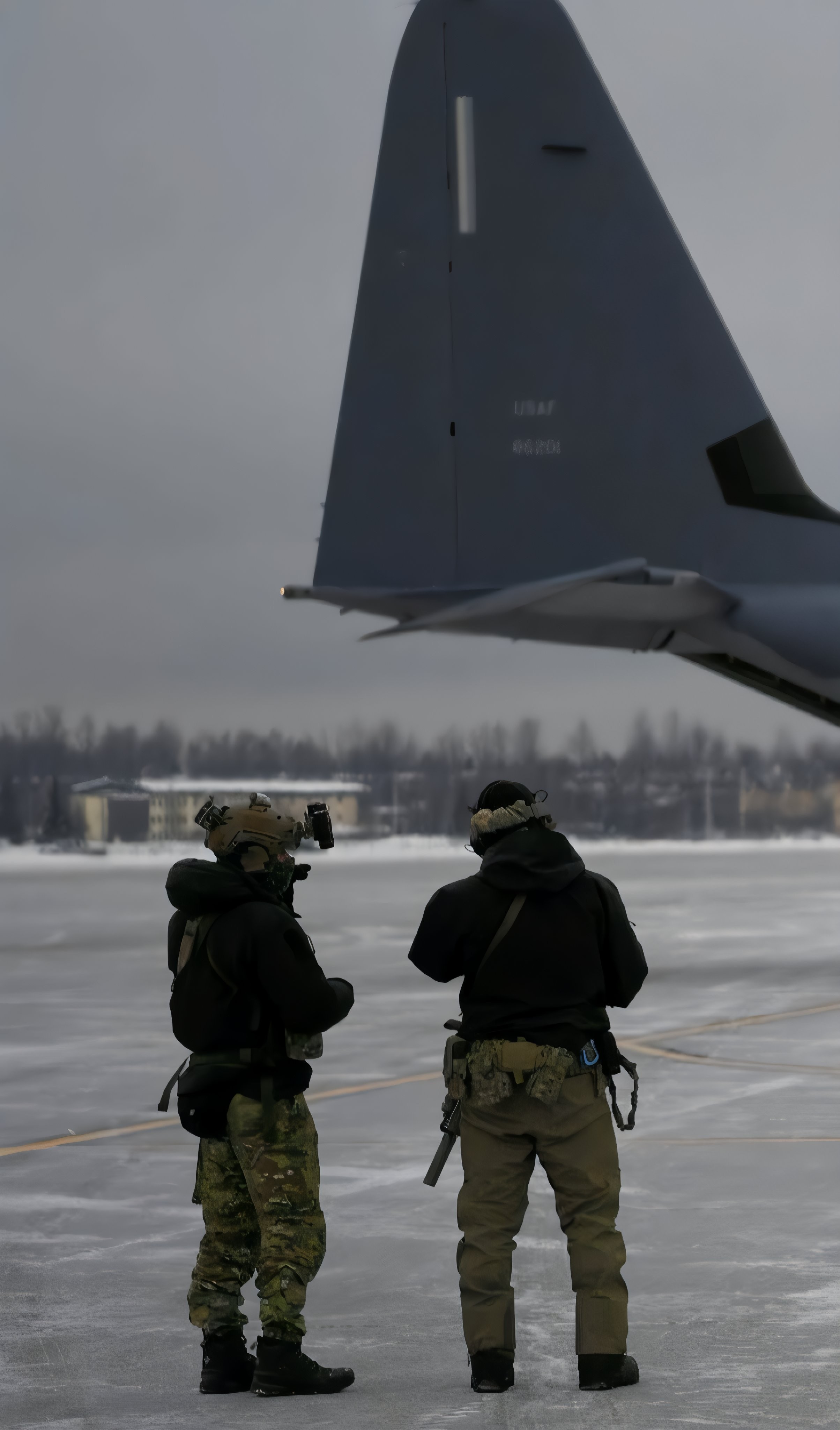二人の完全装備の兵士が雪に覆われた滑走路で大型軍用機の尾部を見上げている
