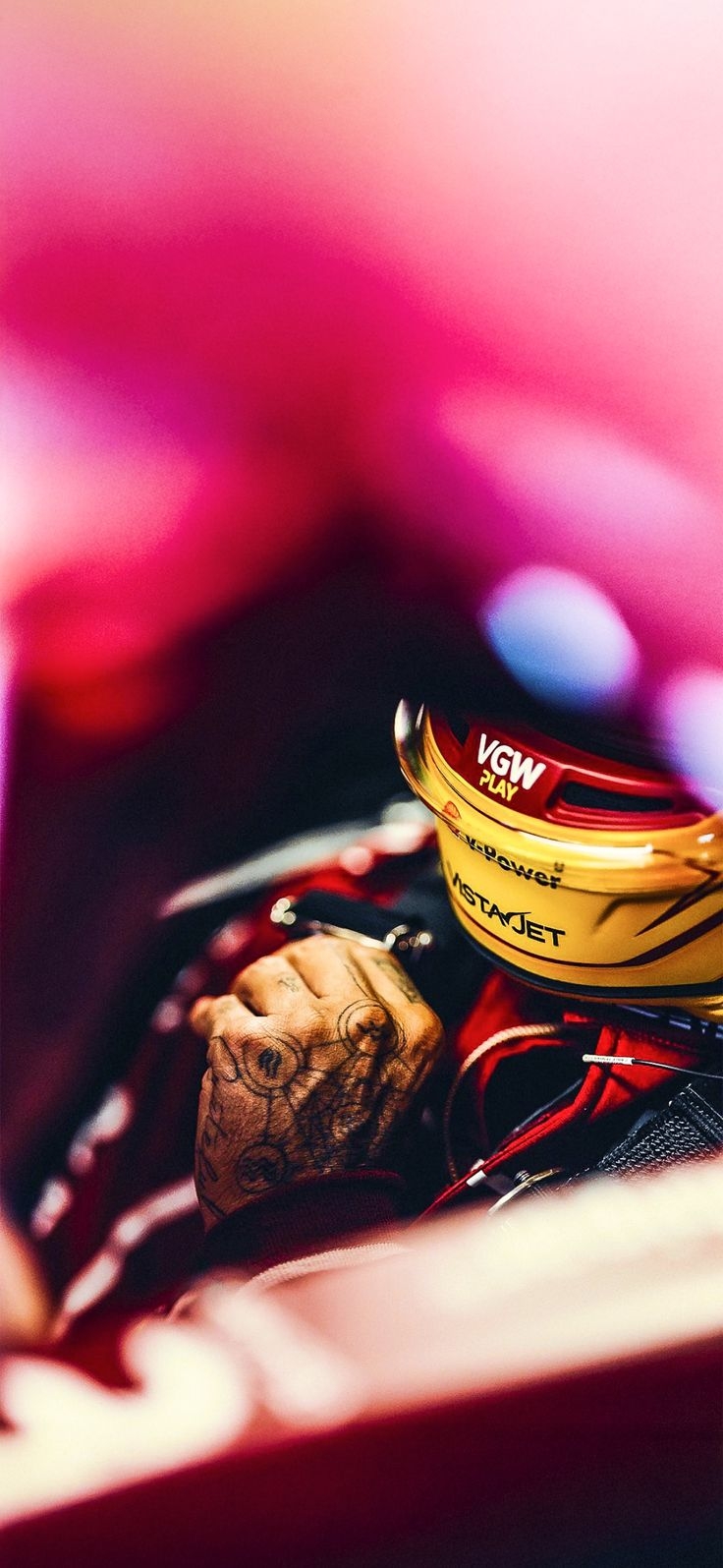 A racer sits in the cockpit wearing a gold helmet and a red racing suit with a pink background
