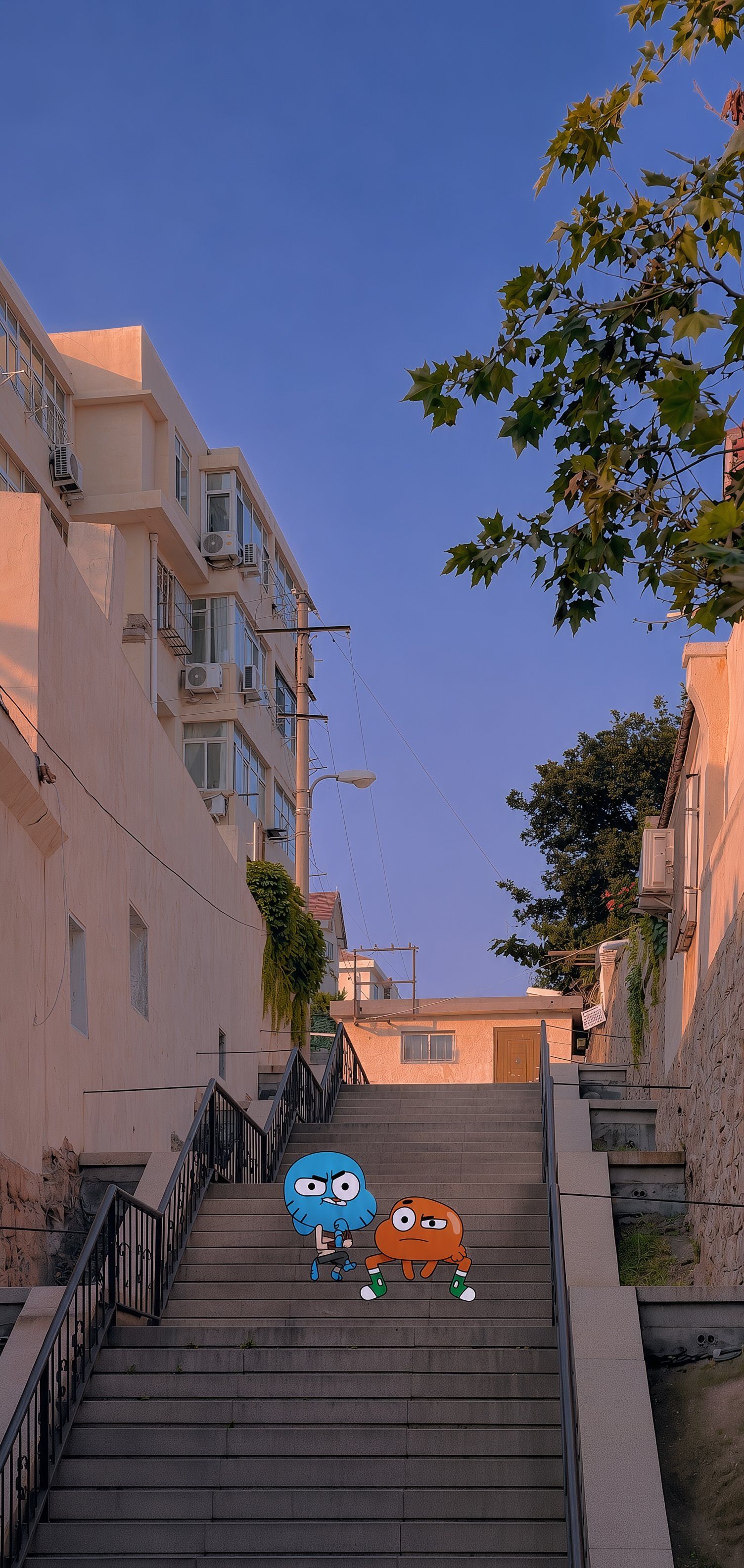 A blue cat and an orange fish sit on a sun-drenched staircase