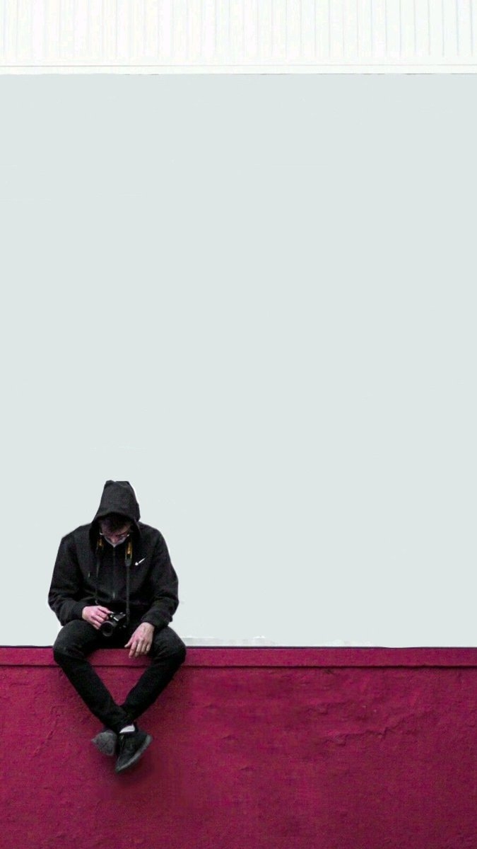 A stark minimalist portrait of a person in black seated against a white wall evoking solitude and introspection