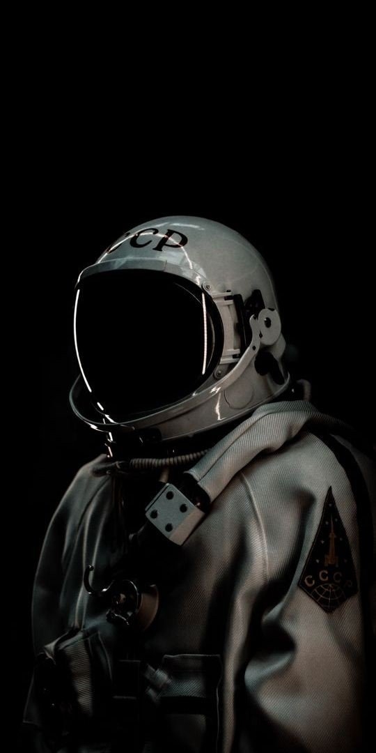 A lone Soviet-era cosmonaut stands in near-total darkness, illuminated by a soft light