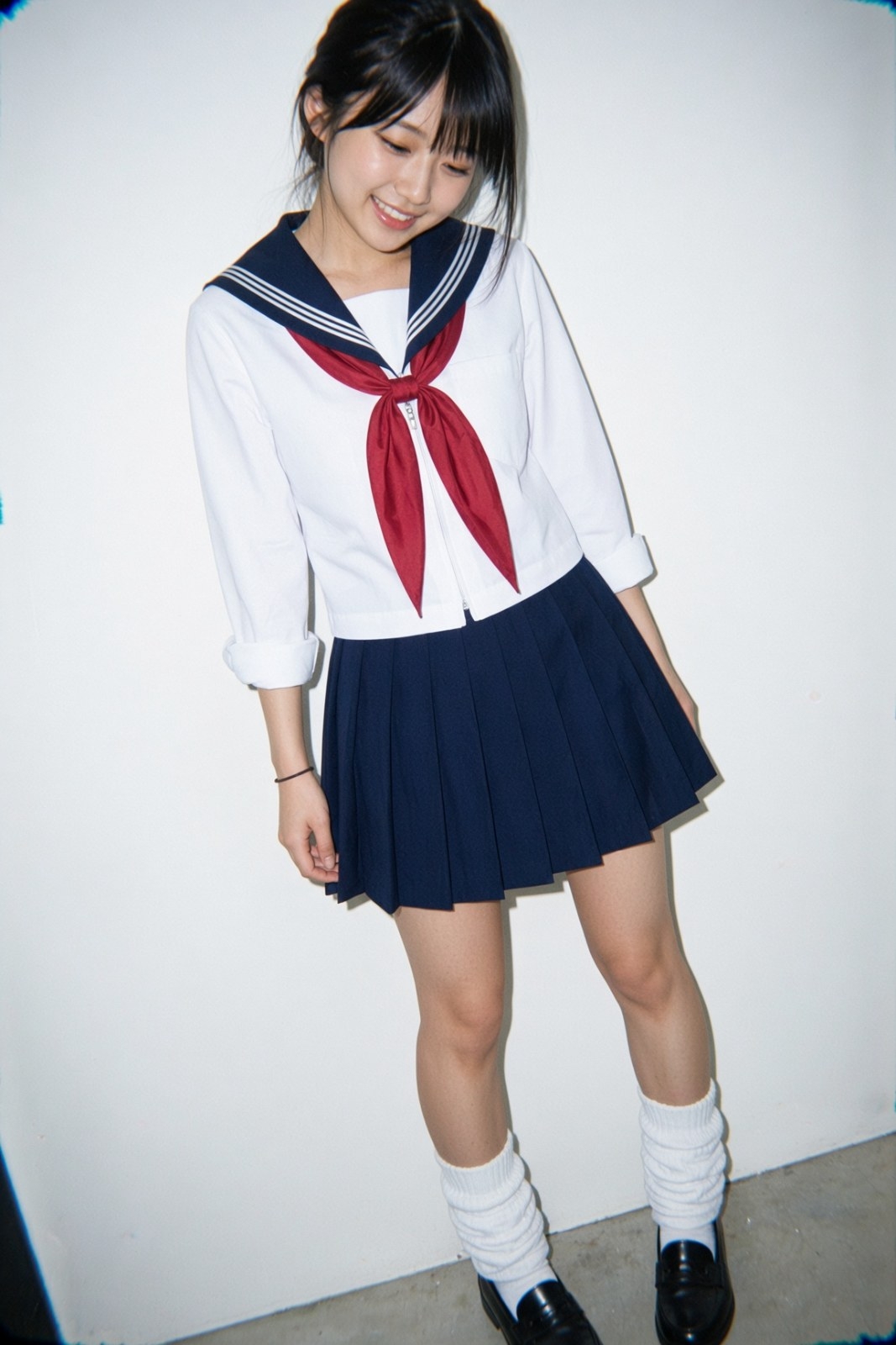 Young woman with long black hair and blunt bangs wears a white sailor top with navy pleated skirt and red scarf before a white wall