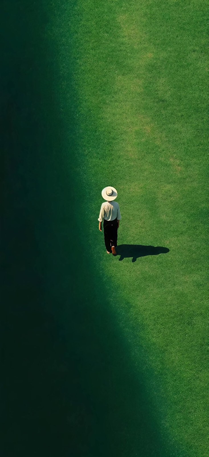Person in wide-brimmed straw hat light top dark pants walks alone on green grass from above